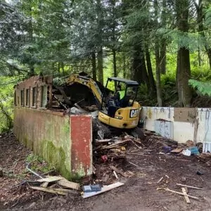 Excavator removing an old wooden structure on a wooded lot